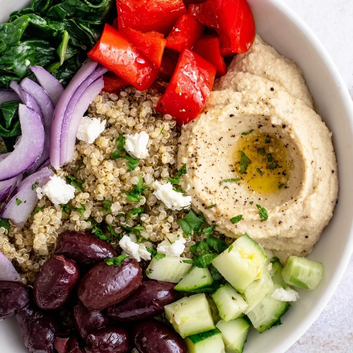 Colorful Mediterranean Quinoa Bowl with feta, olives, and a generous hummus dollop beside sliced cucumbers and tomatoes. 