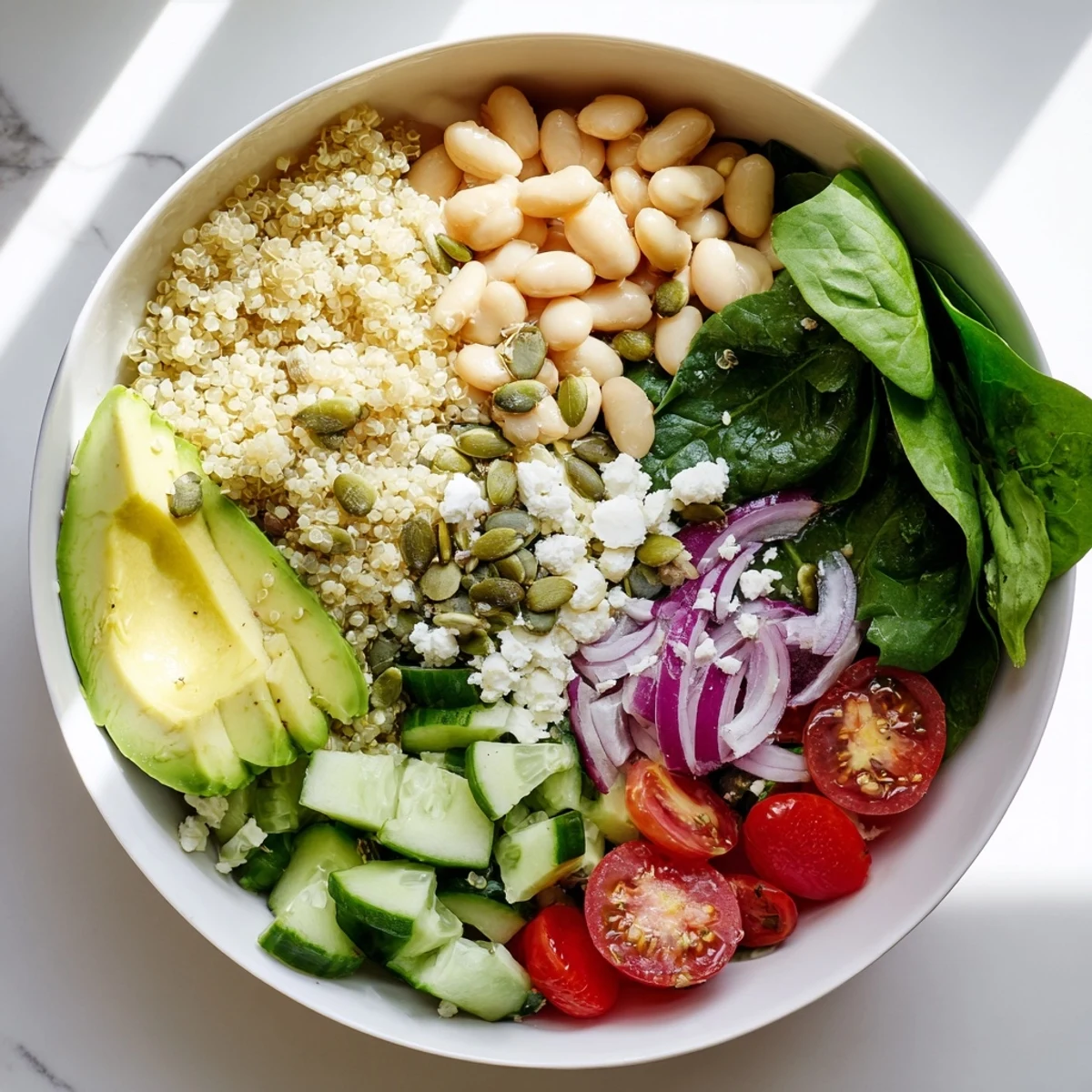 Bright overhead view of a Spinach & White Bean Ultimate Protein Power Bowl with quinoa, spinach, white beans, tomatoes, cucumber, avocado, pepitas, and feta, ready to eat.