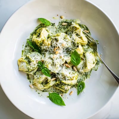 Steaming skillet of cheesy Pesto Alfredo Tortellini tossed in vibrant green basil pesto sauce with visible garlic and pine nut garnish.
